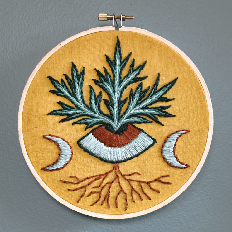 ochre fabric in a wooden hoop embroidered with a mugwort plant blooming up from the center of an eye with a crescent moon on each side
