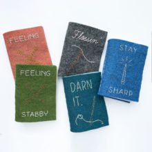 a set of felt needle books in different colors and stitched with images of needles and needle-related puns scattered on a white table