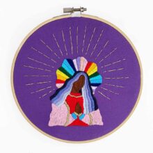 a saint with dark brown skin, blue hair, and a rainbow halo embroidered on purple fabric