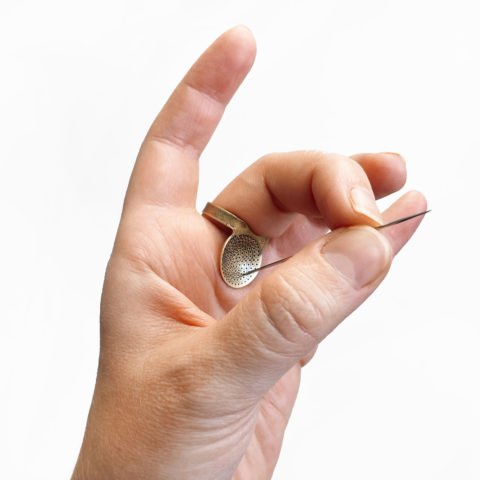 A light-skinned hand holding a long sashiko needle against a brass palm disc sashiko thimble