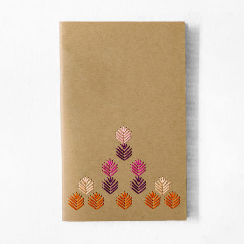 A kraft brown notebook embroidered with a geometric leaf pattern stitched in autumn jewel tones on a white table