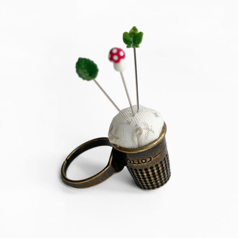 antiqued brass ring with a thimble turned into a tiny pincushion with three decorative straight pins stuck in it