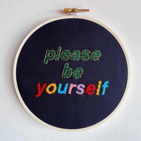 embroidery of colorful letters on black fabric that reads “Please be yourself”