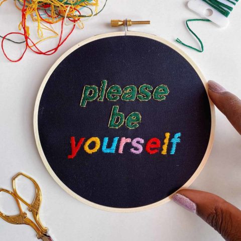 embroidery of colorful letters on black fabric that reads “Please be yourself” in a tiny hoop pinched between two dark-skinned fingers and surrounded by stitching supplies