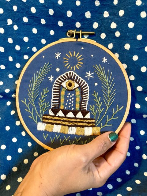 a hand holding royal blue fabric in a wooden embroidery hoop stitched with images of a portal door with an eye on top surrounded by plants and stars in front of a blue fabric with white polka dtos