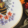 close-up of an embroidery on white of a bee with jewel-shaped thorax, surrounded by flowers and celestial symbols