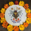 an embroidery on white of a bee with jewel-shaped thorax, surrounded by flowers and celestial symbols inside a wooden hoop and a ring of orange and yellow flowers
