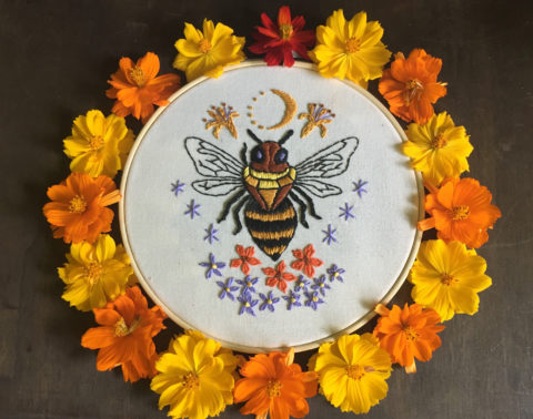 an embroidery on white of a bee with jewel-shaped thorax, surrounded by flowers and celestial symbols inside a wooden hoop and a ring of orange and yellow flowers