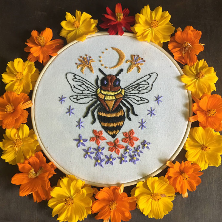 an embroidery on white of a bee with jewel-shaped thorax, surrounded by flowers and celestial symbols inside a wooden hoop and a ring of orange and yellow flowers