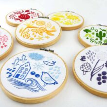 Seven embroidery hoops each showing folk art objects stitched in a different color of the rainbow