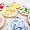 Seven embroidery hoops each showing folk art objects stitched in a different color of the rainbow