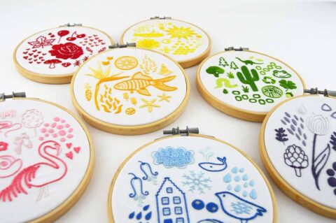 Seven embroidery hoops each showing folk art objects stitched in a different color of the rainbow