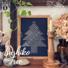 sashiko tree embroidery sampler pattern displayed in a wooden frame on a wooden table surrounded by various decorative objects