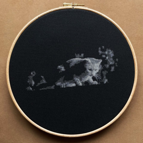 A white cat looking down from a garden fence at night stitched in grayscale on black fabric in a wooden hoop