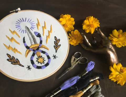 white fabric in a hoop embroidered with a striped snake curled around a shining sowrd with lightning bolts and laurel leaves on either side, laying on a dark wooden table surrounded by embroidery supplies and yellow flowers