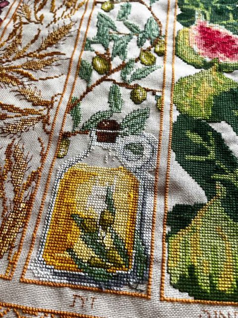 close up of three cross-stitched panels showing barley, olives and oil, and figs