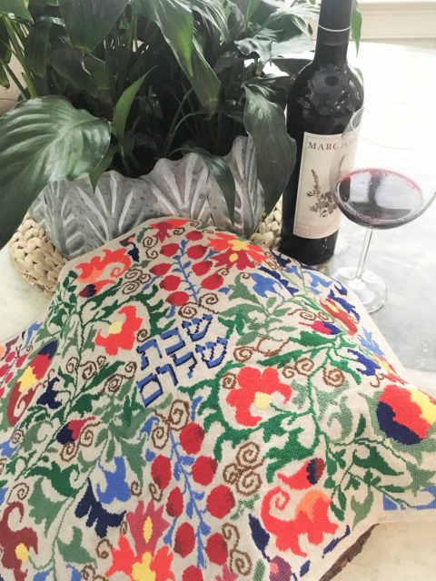 colorful botanical folk art patterns cross-stitched around the hebrew words shabbat shalom on a challah cover displayed next to a potted plant, a wire bottle and a wine glass