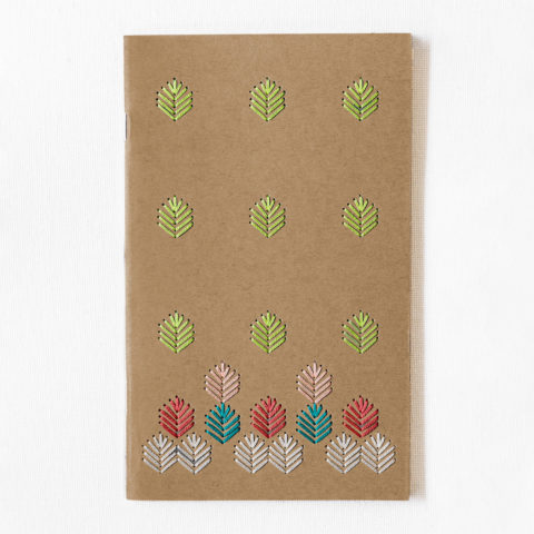 A kraft brown notebook embroidered with a geometric leaf pattern stitched in bright spring colors on a white table