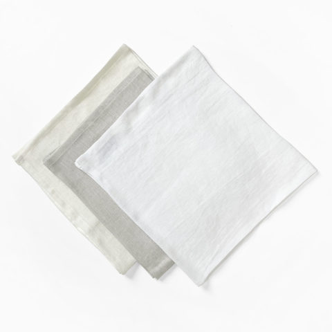 a set of three linen napkins in ivory, light gray, and white, folded into squares and laid in a row