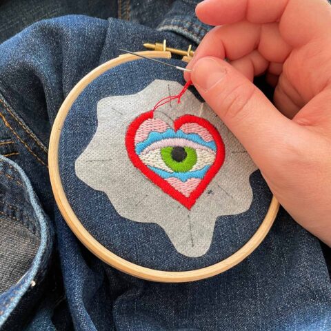 a light-skinned hand embroidering an image of an eye inside a red heart onto a denim jack