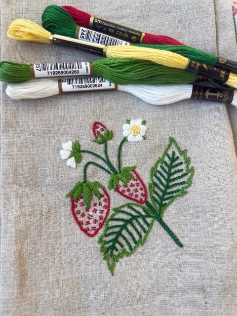 A small spray of strawberries with flowers and leaves embroidered on an ivory linen tea towel displayed next to the threads needed to make it