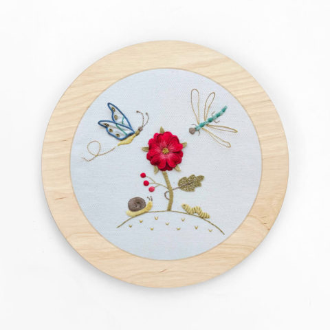 a flower and little garden bugs embroidered on white twill in a circular blond wood frame