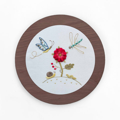a flower and little garden bugs embroidered on white twill in a circular brown wood frame
