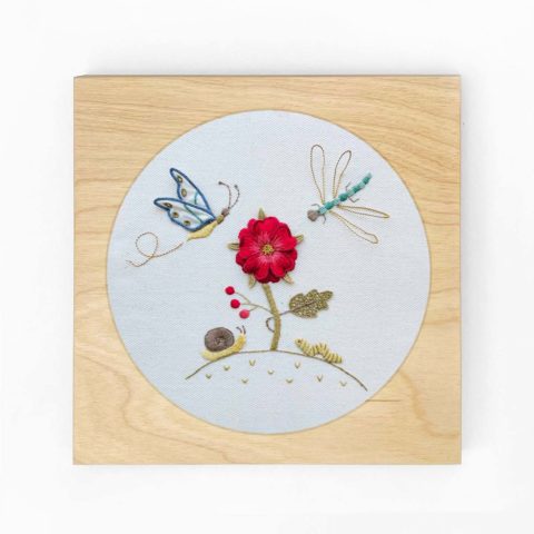 a flower and little garden bugs embroidered on white twill in a square blond wood frame