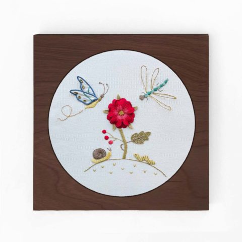 a flower and little garden bugs embroidered on white twill in a square brown wood frame