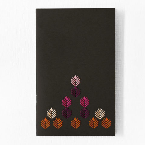A black notebook embroidered with a geometric leaf pattern stitched in autumn jewel tones on a white table