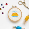 A tiny wooden embroidery hoop showing a cross-stitched taco and the words "tacos fix everything" in blue, laying on a white table surrounded by embroidery supplies and matching rhinestones