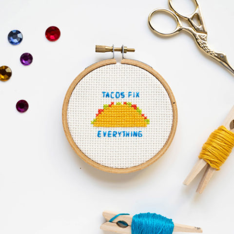 A tiny wooden embroidery hoop showing a cross-stitched taco and the words "tacos fix everything" in blue, laying on a white table surrounded by embroidery supplies and matching rhinestones