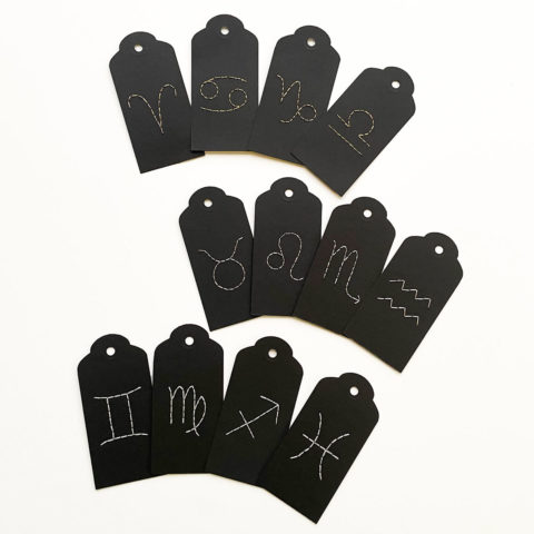 A set of twelve black paper gift tags embroidered with white Western zodiac symbols