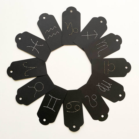 A set of twelve black paper gift tags embroidered with white Western zodiac symbols arranged in a ring