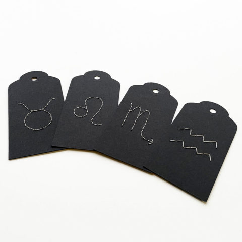A set of four black paper gift tags embroidered with white Western zodiac symbols