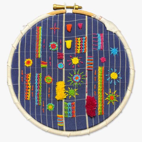 An embroidery stitch sampler of small geometric designs in bright primary colors on dark blue fabric
