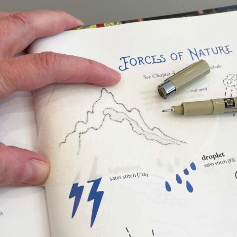 a hand and a pen tracing a mountain shape from a book onto translucent paper