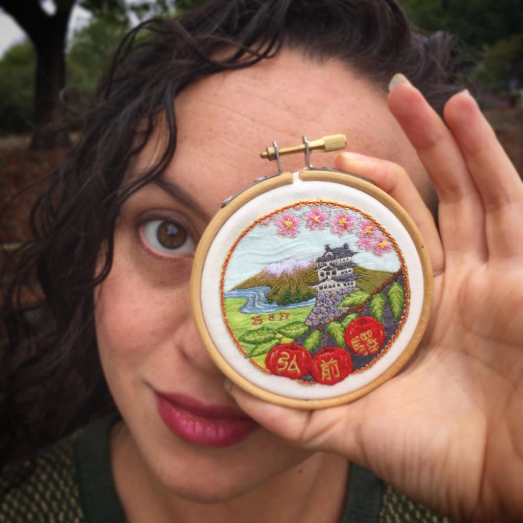 embroidery artist yuka hoshino of mayuka fiber art holding a small hoop in front of her eye showing an embroidery of a japanese landscape with pagoda, cherry blossoms, and red lanterns