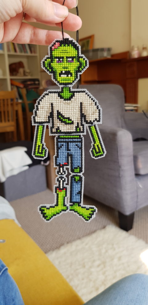light-skinned hand holding a zombie cross stitch hanging decoration